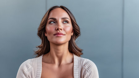 A woman with shoulder-length hair sits quietly, her expression thoughtful as she looks upwards. The backdrop features a sleek gray wall, creating a calm atmosphere.の素材