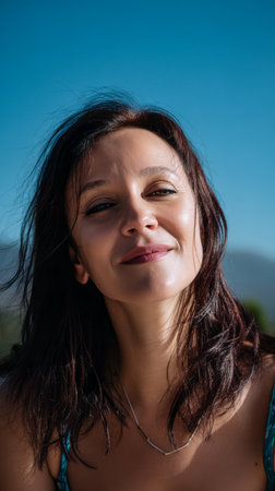 A woman enjoys the warm sunshine with a bright smile on her face. The clear blue sky and distant mountains create a peaceful outdoor setting. It is a joyful moment captured in nature.の素材