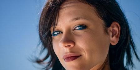 A woman with striking blue eyeliner smiles against a clear blue sky. The sun shines bright, illuminating her joyful expression as she enjoys a relaxing beach day.の素材