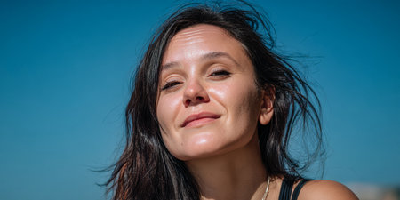 A woman is standing outdoors, smiling gently as the sun shines brightly. Her dark hair flows in the breeze under a clear blue sky, creating a relaxing atmosphere.の素材