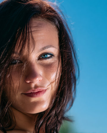 A woman with striking blue eyes and messy hair smiles warmly at the camera. The setting is bright and sunny with a clear blue sky in the background, creating a cheerful atmosphere.の素材