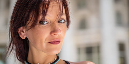 A woman with short dark hair stands outdoors, smiling and looking directly at the camera. Bright sunlight highlights her features and the urban landscape behind her.の素材
