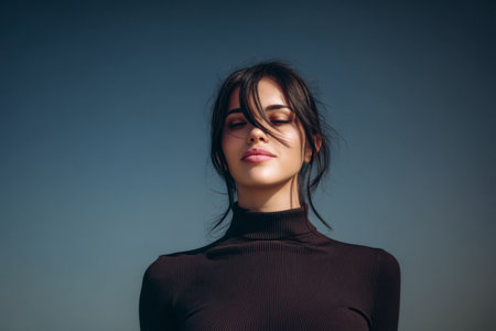 A young woman stands outdoors, basking in sunlight with her hair gently blowing in the wind. She exudes calm and confidence against a backdrop of clear blue sky.の素材