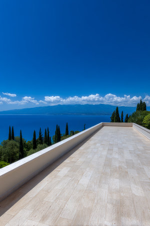 A bright terrace offers a breathtaking view of calm blue waters and lush green trees under a clear blue sky. Distant mountains can be seen on the horizon, enhancing the tranquil scene.の素材