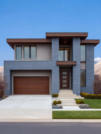 A modern house features sleek architecture with a blend of brick and wood elements. The driveway leads to a stylish entrance, surrounded by well-kept landscaping and mountains in the background.の素材