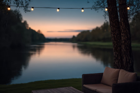Soft lights hang above a peaceful riverbank as evening falls. A stylish sofa and table invite relaxation while the sky changes colors over the calm water.の素材
