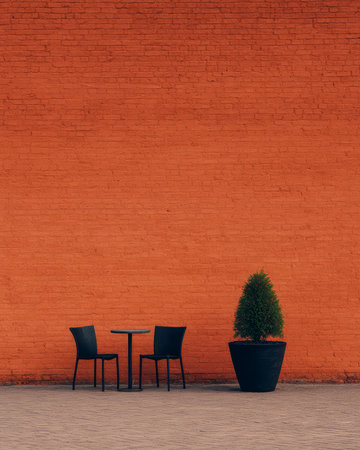 Two black chairs and a small table sit in a cozy outdoor setting beside a green potted plant, all situated against a bright orange brick wall on a sunny day.の素材