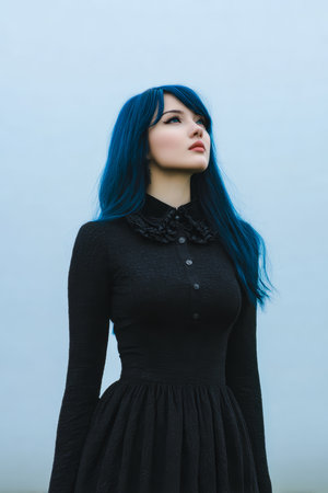 A young woman stands outdoors on a misty day, looking up thoughtfully at the cloudy sky. She wears a black dress and has striking blue hair, creating a dramatic contrast.の素材