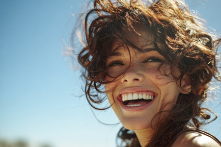 A woman laughs joyfully as her hair blows in the wind. She enjoys a sunny day outdoors, surrounded by a clear blue sky and warm atmosphere, smiling brightly.の素材