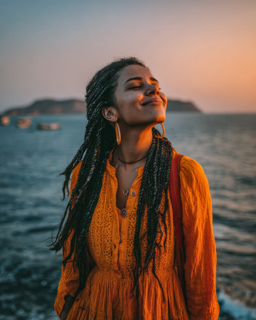 A woman stands by the water, dressed in a bright orange dress. She smiles softly as the sun sets, reflecting golden hues on the ocean waves and hills nearby.の素材