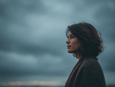 A woman stands quietly, looking at a cloudy sky at dusk. Her thoughtful expression reflects the serene yet dramatic atmosphere along the shoreline.の素材