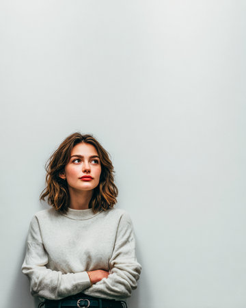 A young woman with shoulder-length hair stands against a light gray wall, wearing a soft sweater. She appears deep in thought, reflecting on her surroundings.の素材