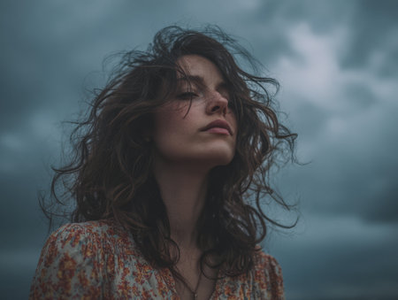 A woman stands outdoors with her curly hair blowing in the wind. She looks thoughtfully towards the horizon, surrounded by a dramatic sky filled with dark clouds during dusk.の素材