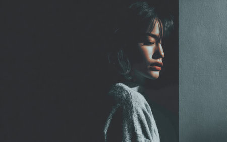 A woman stands quietly in a low-light setting, her features partially obscured by soft shadows. She appears thoughtful, lost in her own reflections as the light gently highlights her profile.の素材