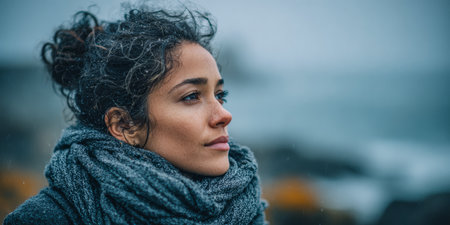 A woman stands by the ocean, her curly hair slightly tousled by the wind. She wears a warm scarf and gazes thoughtfully at the waves under a gray sky filled with clouds, conveying a sense of calm.の素材