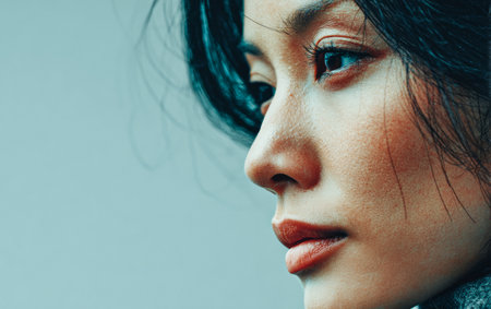 A young woman with dark hair and delicate features shows a contemplative expression against a neutral background. Natural light enhances her serene appearance and relaxed mood.の素材