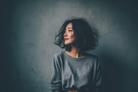 A woman with wavy hair smiles softly while posing against a textured gray wall. Her casual sweater highlights her relaxed demeanor in a serene indoor setting.の素材