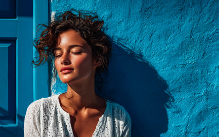 A young woman stands by a bright blue wall, eyes closed, enjoying the warmth of the sun on her face. Her curly hair catches the light, creating a tranquil and thoughtful mood.の素材