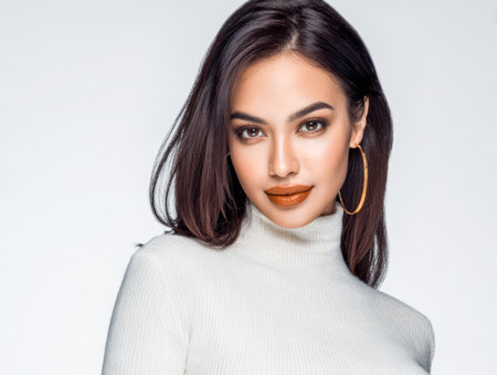 A woman with long dark hair smiles confidently while wearing a white sweater. She has striking makeup and golden hoop earrings, showcasing her fashionable style and elegance.の素材