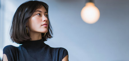 A woman with short hair shows a serious expression as she stands near a light fixture. She appears deep in thought while dressed casually in a soft setting.の素材