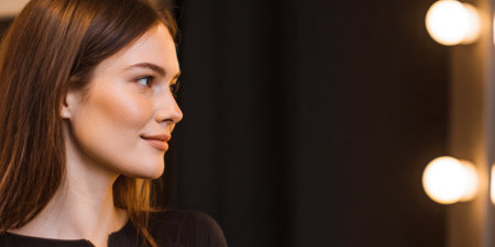A woman is seated in a salon chair, looking to the side while sitting near a mirror. Bright lights illuminate the space, creating a focus on her profile.の素材