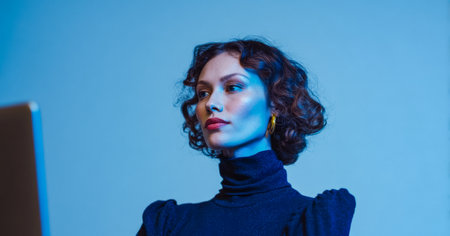 A woman with curly hair stands in front of a laptop. She is focused on the screen while the background features blue lighting. The setting appears to be indoors.の素材