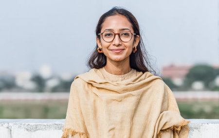 A young woman stands on a rooftop, smiling gently. She wears simple clothing and has long hair. The city and clear sky are visible in the background during daytime.の素材