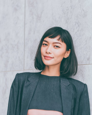 A woman stands against a gray wall with a smile. She has short hair and wears a dark outfit. The setting is bright and modern. There is a hint of urban style around her.の素材