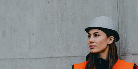 Woman wearing a safety helmet and vest stands near a concrete wall at a construction site. She looks thoughtfully to the side, focused on her work.の素材
