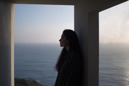 A woman leans against a wall near a window. She looks out at the ocean. The water appears calm and the sky is clear. It is daytime and sunlight is present.の素材
