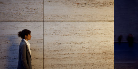 A woman stands next to a large stone wall in an urban area during the evening. The setting has low light, creating shadows and highlighting the texture of the wall.の素材