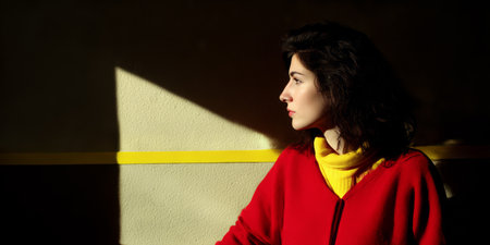 A woman sits alone in a room with sunlight casting shadows on the wall. She gazes thoughtfully to the side. The setting features a yellow stripe and soft light.の素材