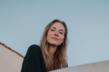 A woman with long hair smiles while leaning on a low wall. The background shows a clear blue sky and part of a building roof. The moment captures a serene atmosphere.の素材