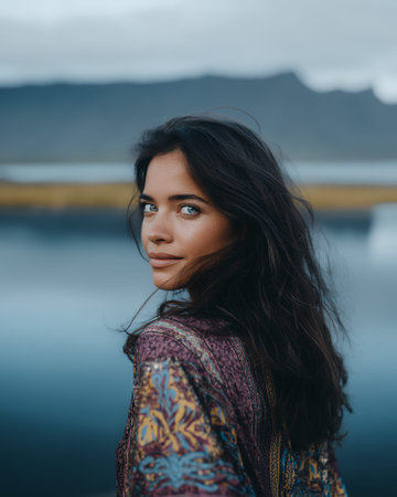 A young woman with dark hair and striking blue eyes looks over her shoulder, her gaze direct and captivating. She is wearing a colorful, patterned garment, possibly a dress or shawl, with intricate designs in shades of purple, yellow, and blue. The background is a soft blur of muted blues and grays, suggesting a natural landscape with water and distant hills or mountains under an overcast sky. The overall mood is serene and contemplative, with the woman's expression hinting at a moment of quiet reflection or connection with her surroundings.の素材