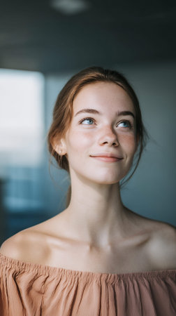 A young woman with freckles and blue eyes looks up and to the side with a slight smile, her hair pulled back and wearing an off-the-shoulder top.の素材