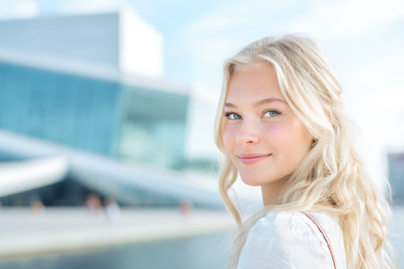 A young woman with blonde hair and blue eyes smiles at the camera in front of a modern building.の素材