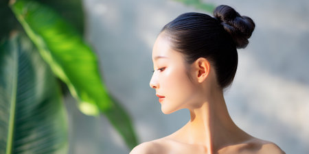 A young Asian woman with her hair in a bun, looking to the side with soft lighting on her face and neck, with a blurred green leaf in the foreground.の素材
