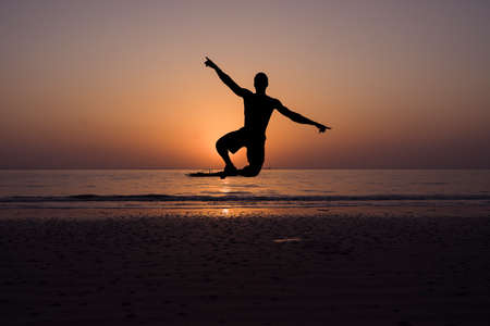 Man on the beach at sunset against light jumping. His arms are stretched out and his legs are bent, touching each other's feet. It's on the beach by the sea.の写真素材