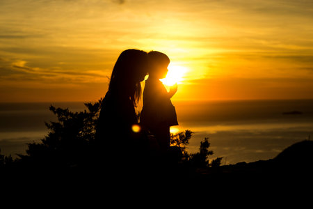 Mother with long hair and small daughter standing, in profile, back at sunset.の写真素材