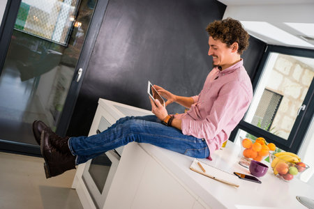 A young white man with curly, smiling hair in jeans and a pink shirt sitting on the kitchen counter, looking at the tablet while drinking coffee in a purple cup. The kitchen is all white.の写真素材