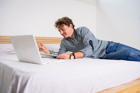 White man with blue plaid shirt and jeans, lying in bed with white sheets and wooden headboard, with...