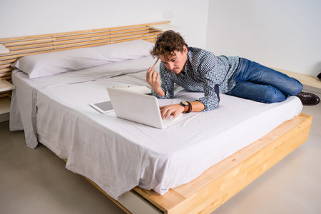 White man with blue plaid shirt and jeans, lying in bed with white sheets and wooden headboard, with computer and mobile phone. She scratches her hair with the pen in a thoughtful way.の写真素材