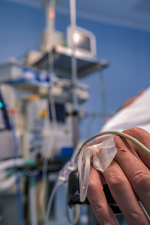 Patient's hand with a serum and an oxygen saturation meter on the finger. Monitor and respirator with all the wires in the back.の写真素材