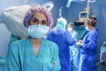 Surgery assistant looking at the camera with the surgery team working in the background. He's wearing a purple hat.の写真素材