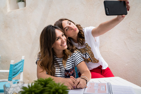 2 girls making a selfie on a terrace. They are laughing and making faces. They are elegant but informal. Their hair is long and wavy.の写真素材