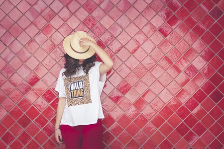 Girl posing with a hat in front of a red wall. She has the hat in her hand. She wears red pants and a white t-shirt. Horizontal photo with space on the rightの写真素材