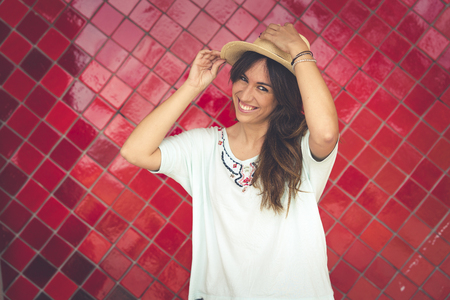 Girl posing with a hat in front of a red wall. She has the hat in her hand. She wears red pants and a white t-shirt. Horizontal photo with space on the rightの写真素材