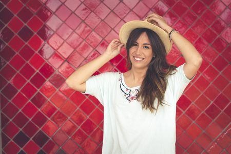Girl posing with a hat in front of a red wall. She has the hat in her hand. She wears red pants and a white t-shirt. Horizontal photo with space on the rightの写真素材