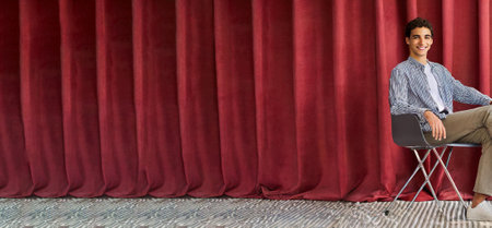 Confident young male in trendy casual outfit sitting with red curtain in the background with copy spaceの素材