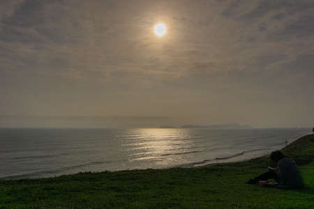 Sunset Landscape in the shore of Lima Peru, silhouette of girl reading, grassの写真素材